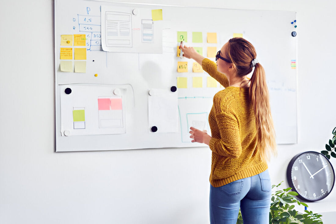 A woman in a yellow sweater and blue jeans brainstorms ideas on a dry erase whiteboard